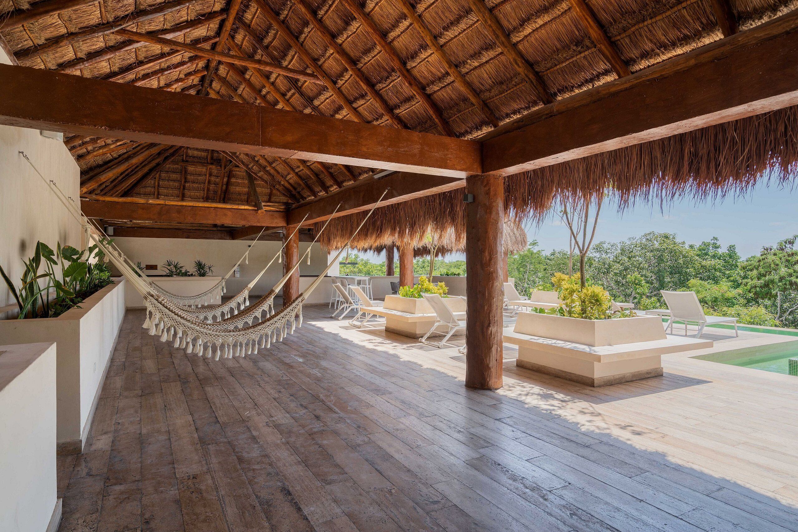 Un espacio al aire libre cubierto con suelo de madera, una hamaca, sillas de descanso y macetas con plantas.