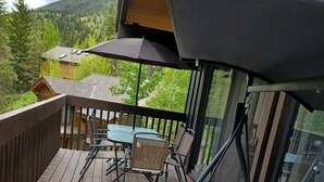 Una terraza de madera con una mesa y sillas, barandilla de vidrio y vistas a una montaña boscosa.