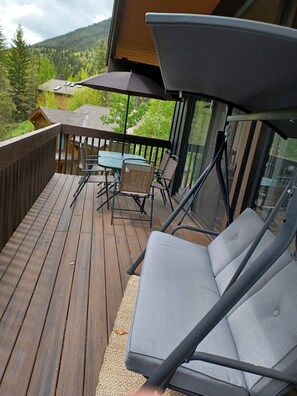 Eine Holzterrasse mit Tisch und Stühlen, einem Glasgeländer und Blick auf einen bewaldeten Berg.