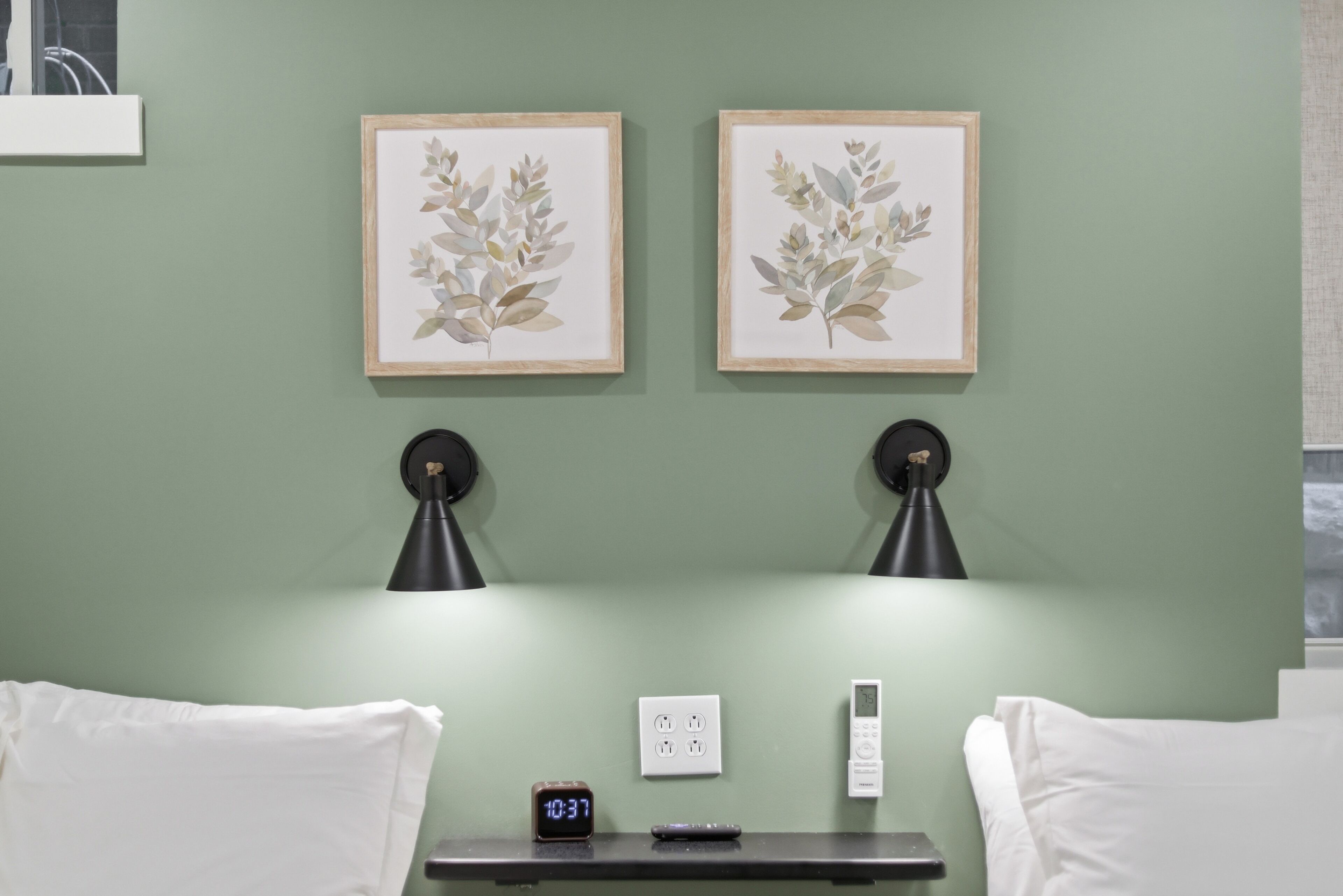 Une chambre d’hôtel avec deux reproductions botaniques encadrées accrochées au mur, deux lampes fixées au mur, une table de chevet avec une horloge et une télécommande, et deux serviettes blanches.