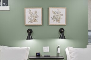 Habitación de hotel con dos láminas botánicas enmarcadas en la pared, dos lámparas de pared, una mesita de noche con un reloj y un control remoto, y dos toallas blancas.