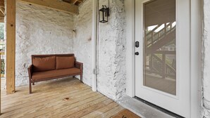 A covered porch with a wooden bench, a white door with a security screen, and a wall with exposed brick.