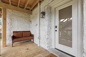 A covered porch with a wooden bench, a white door with a security screen, and a wall with exposed brick.