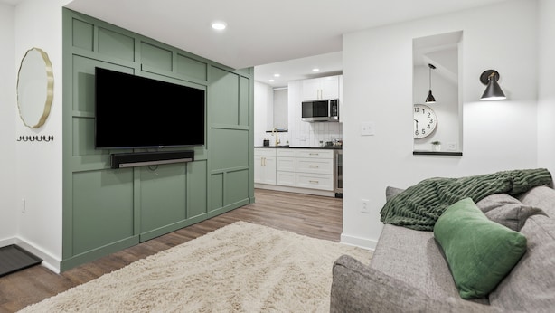 A modern living room with a flat-screen TV, a green accent wall, a white sofa with a green throw, and a kitchen in the background.