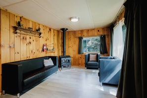 Una habitación con interior de madera, un sofá negro, un sillón azul, una chimenea y una ventana con cortinas.
