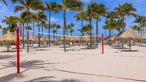Cancha de voleibol de playa con red, palmeras y sombrillas de paja.