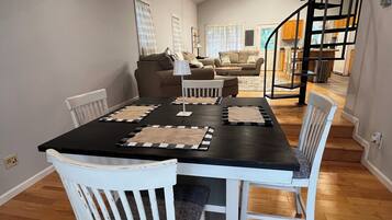 Un coin repas avec une table noire et des chaises blanches, un escalier et, au fond, un salon.