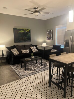Ein modernes Wohnzimmer mit schwarzem Sofa, Couchtisch, Esstisch und Deckenventilator.