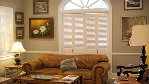 A living room with a brown leather sofa, a coffee table, a lamp, and a window with shutters. - Comfort and Style, Modern Corporate Apartment in Pascagoula (Pascagoula)