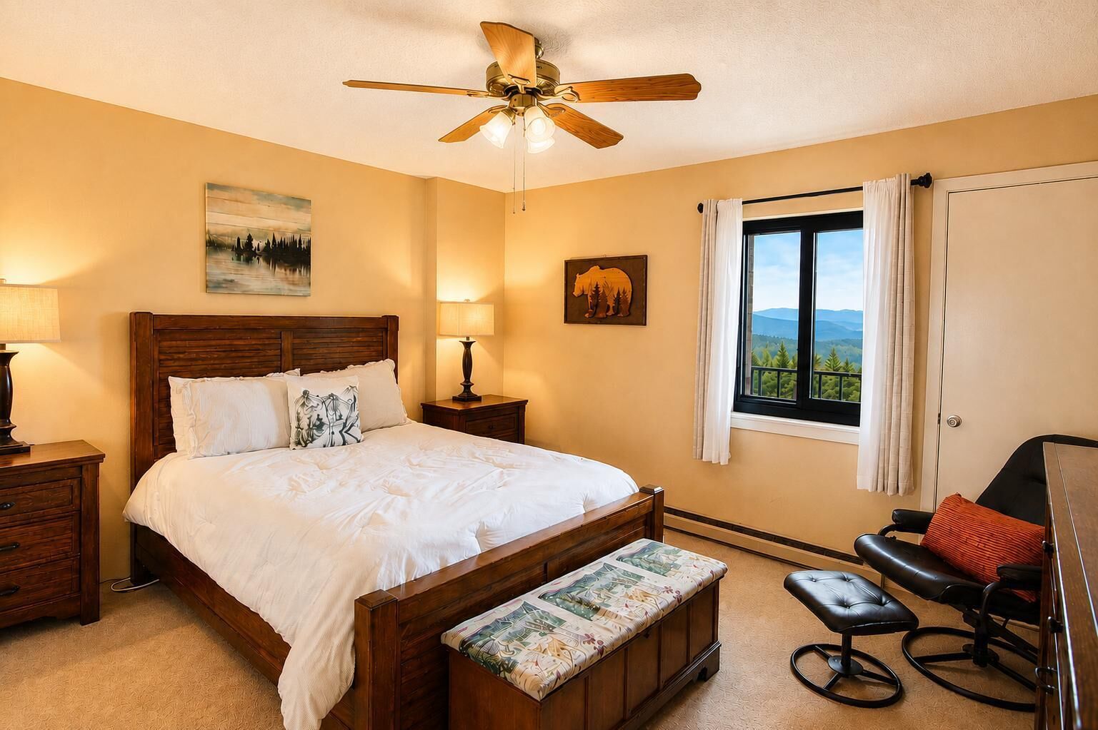 A bedroom with a wooden bed, bedside lamps, a desk, a chair, a window with a view, and a ceiling fan.