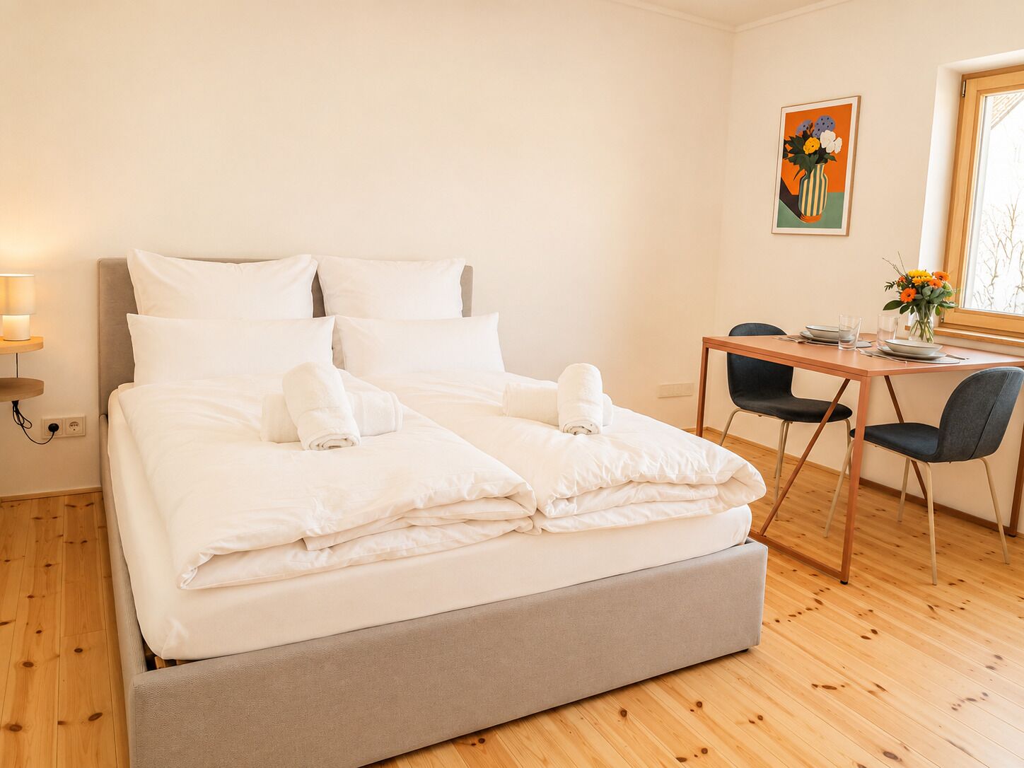 A neatly made bed with white linens, a bedside table with a lamp, a small table with a vase of flowers, and a chair.