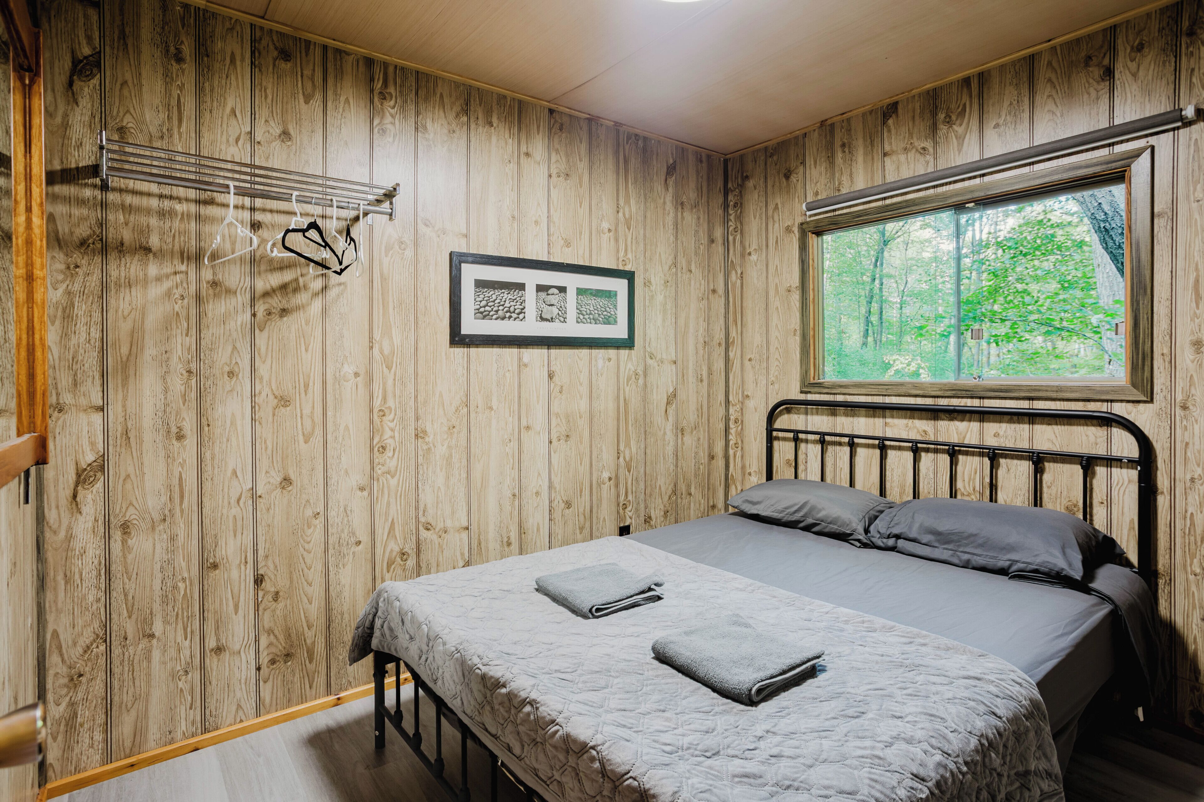 Une chambre avec un lambris en bois, un cadre de lit en métal, une fenêtre donnant sur des arbres et un tableau encadré au mur.