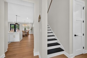 Un intérieur moderne avec un escalier menant à un deuxième étage, un coin repas agrémenté d’un lustre, et une cuisine dotée de meubles blancs.