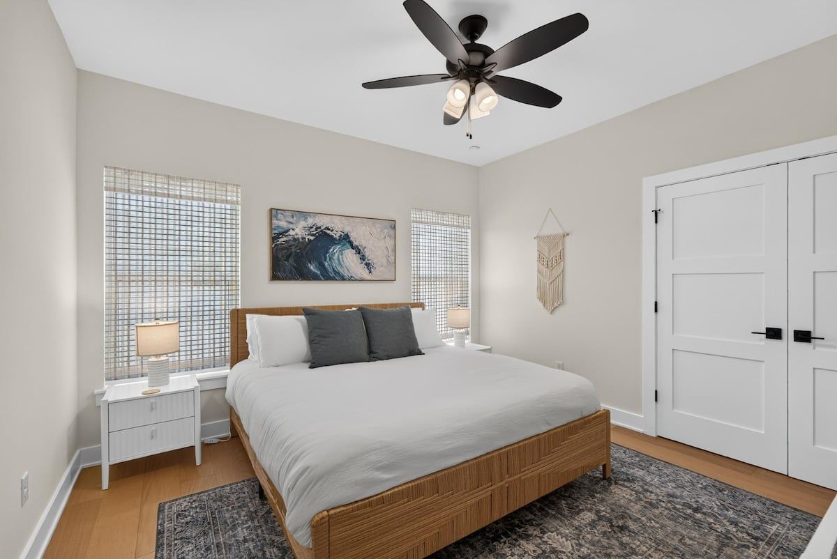 Une chambre à coucher comprenant un lit, des tables de chevet, un ventilateur de plafond, une fenêtre avec des stores, une œuvre d’art accrochée au mur et une armoire.