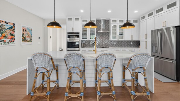 Una cocina moderna con armarios blancos, una isla central con taburetes altos, iluminación colgante y un refrigerador de acero inoxidable.