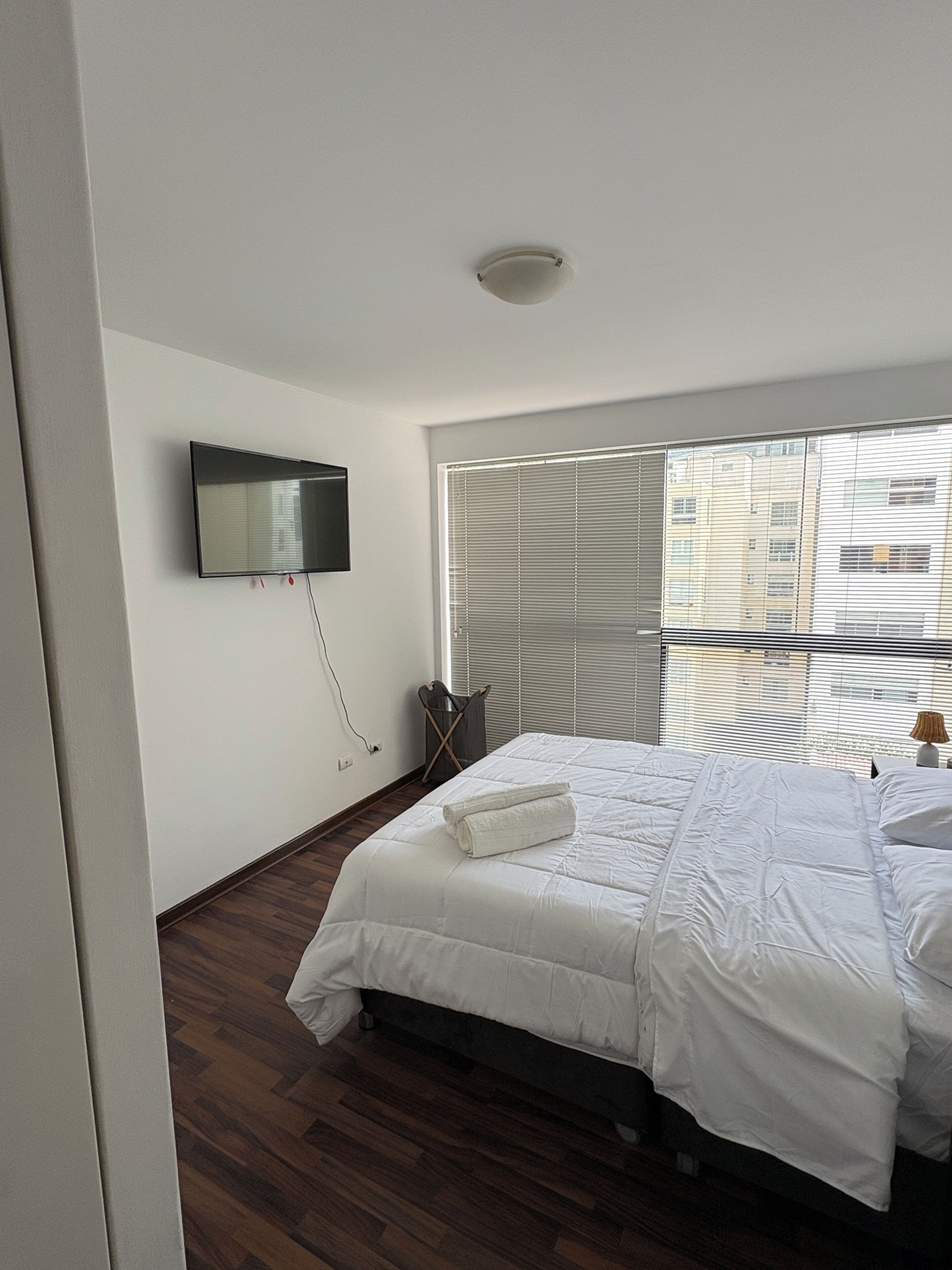 A bedroom with a bed, a TV mounted on the wall, a window with blinds, and a ceiling light.