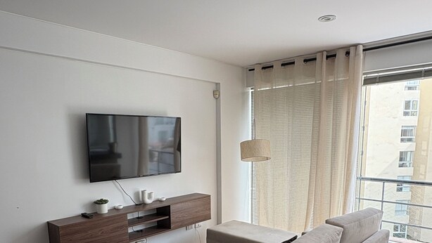 Ein modernes Wohnzimmer mit Flachbildfernseher, Holzboden, einer Couch, einem kleinen Couchtisch und einem großen Fenster mit Vorhängen.