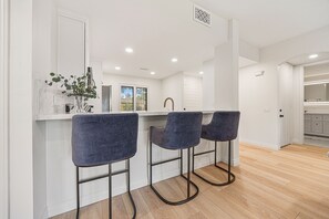 A modern kitchen with a bar area, featuring three bar stools, a marble countertop, and a view of the outdoors. - Your Perfect Desert Retreat -Relax and Recharge in a fully remodeled 2 bed condo (Palm Springs)