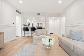 A modern living room with a sofa, a coffee table with flowers, and a kitchen in the background. - Your Perfect Desert Retreat -Relax and Recharge in a fully remodeled 2 bed condo (Palm Springs)