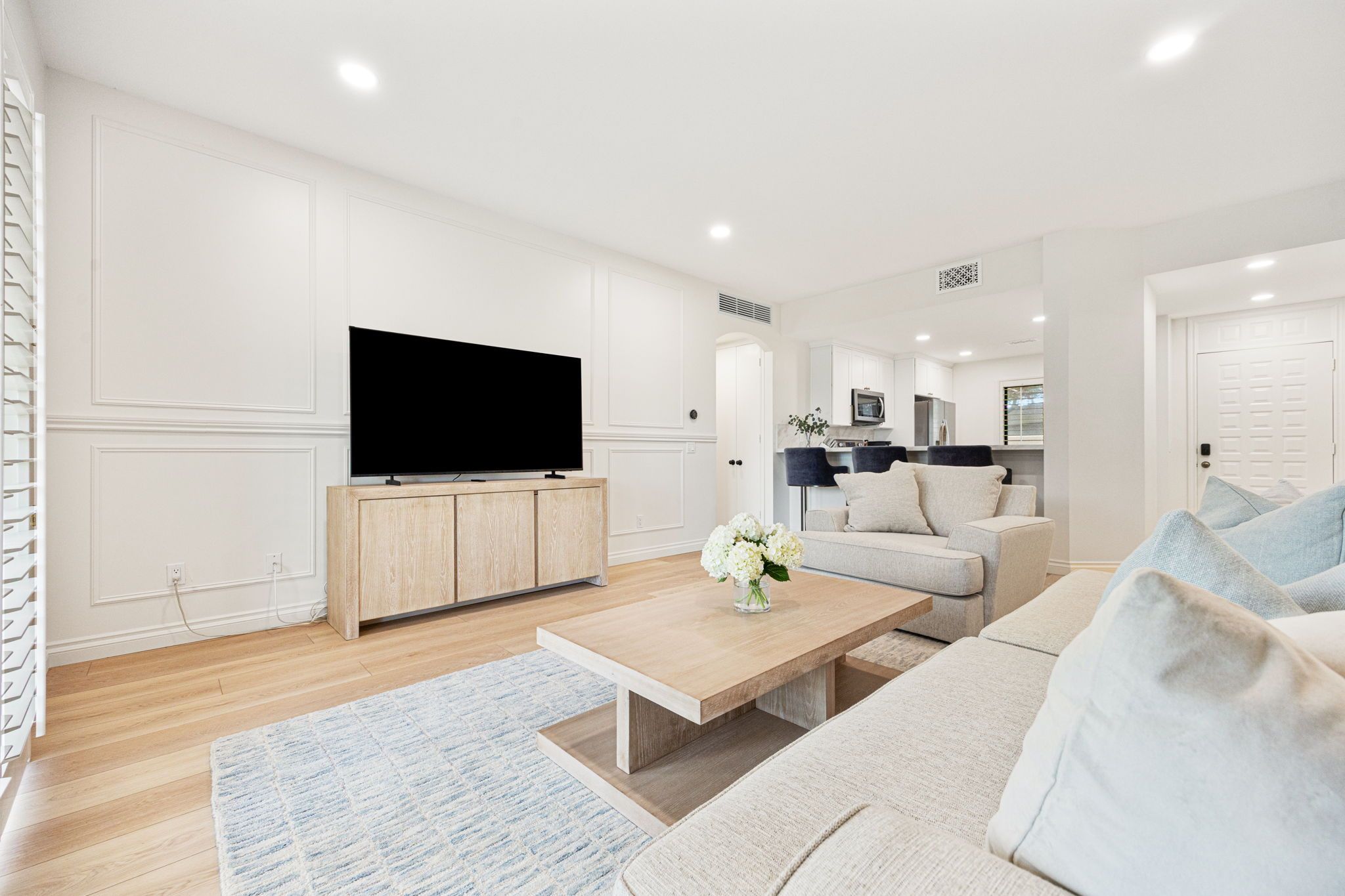 A modern living room with a flat-screen TV, a wooden cabinet, a light-colored sofa, a coffee table, and a kitchen area in the background.