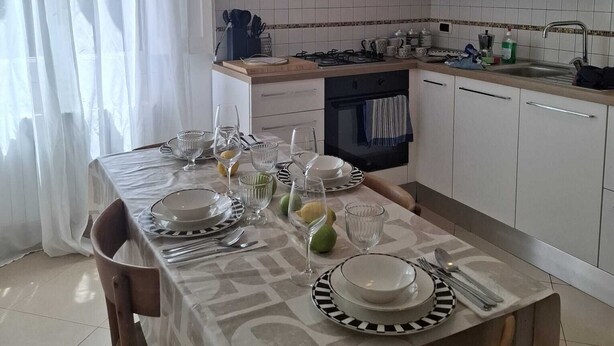 Ein Essbereich mit Tisch für vier Personen, eine Küche im Hintergrund und ein Fenster mit durchsichtigen Vorhängen.
