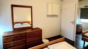 Et soveværelse med en trækommode, en seng med et mønstret sengetæppe, et spejl, et hvidt skab og en dør med en udgangsskilte.
