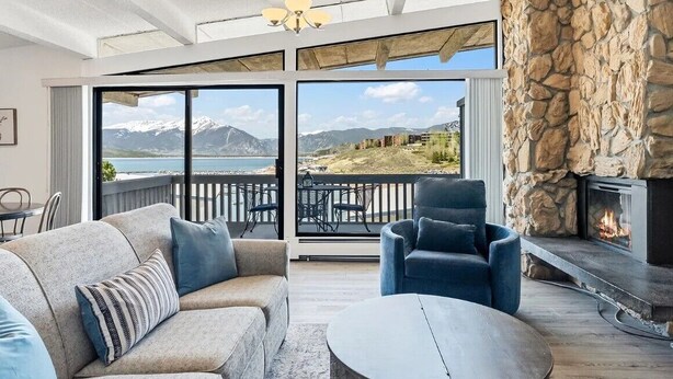 Una sala de estar moderna con chimenea de piedra, un sofá, una silla y vistas a un paisaje montañoso a través de grandes ventanales.
