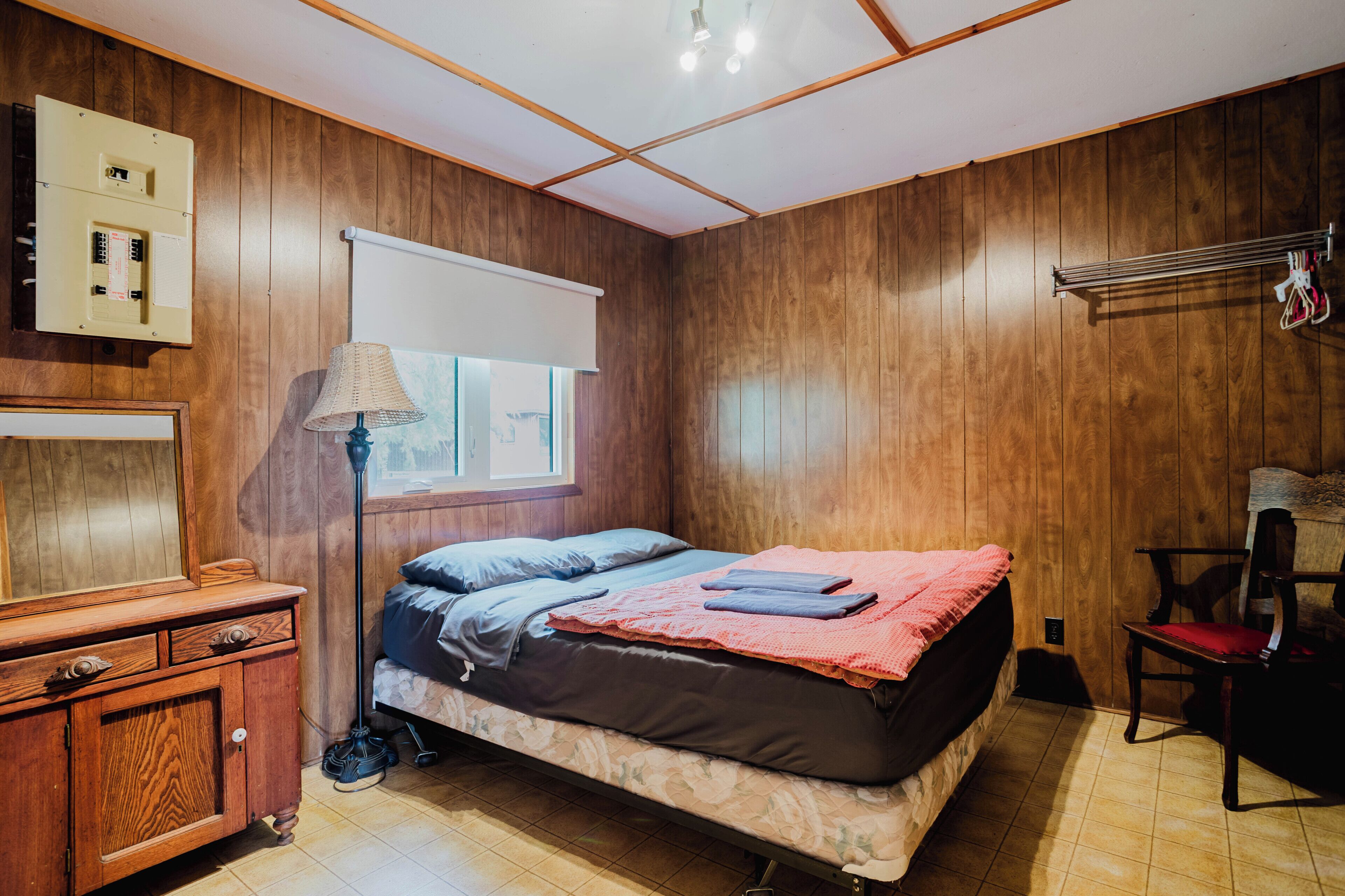 A bedroom with a bed, wooden walls, a window, a lamp, a chair, and a cabinet.