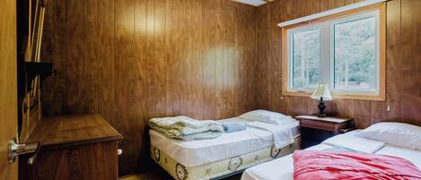 A room with wooden walls, two beds, a small table with a lamp, and a window with a view of trees.