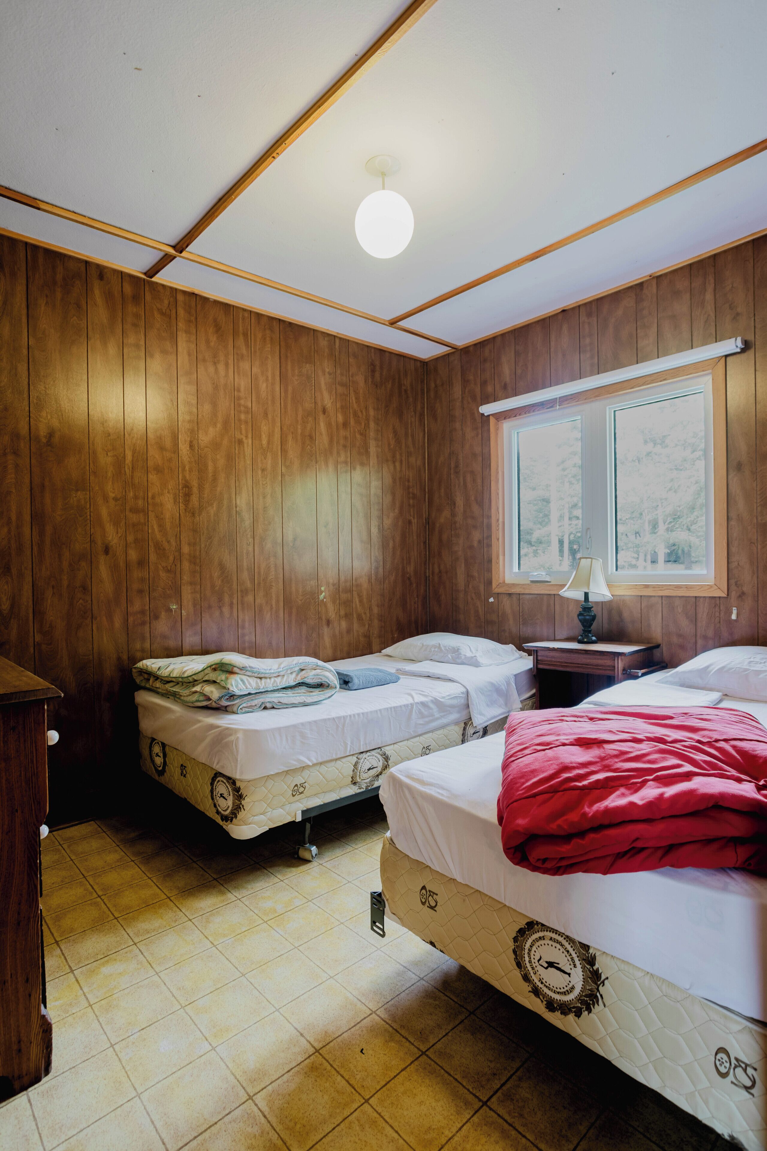 Ein Zimmer mit zwei Betten, Holzwänden, einem Fenster mit Blick und einer Deckenleuchte.
