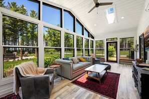 A spacious room with a large window, a leather sofa, a coffee table, and a dining area. - Modern Rustric Retreat w/Fenced Yard + 4-Season Room (Baileys Harbor)