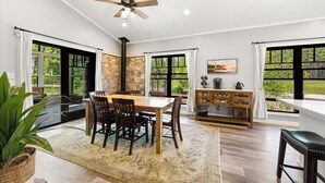 A dining area with a wooden table and chairs, a ceiling fan, and large windows with curtains. - Modern Rustric Retreat w/Fenced Yard + 4-Season Room (Baileys Harbor)