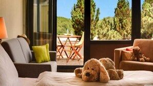 A room with a bed, a sofa, a teddy bear, and a view of trees through large windows. - 2 Person Holiday Park Home in Willingen (Willingen)