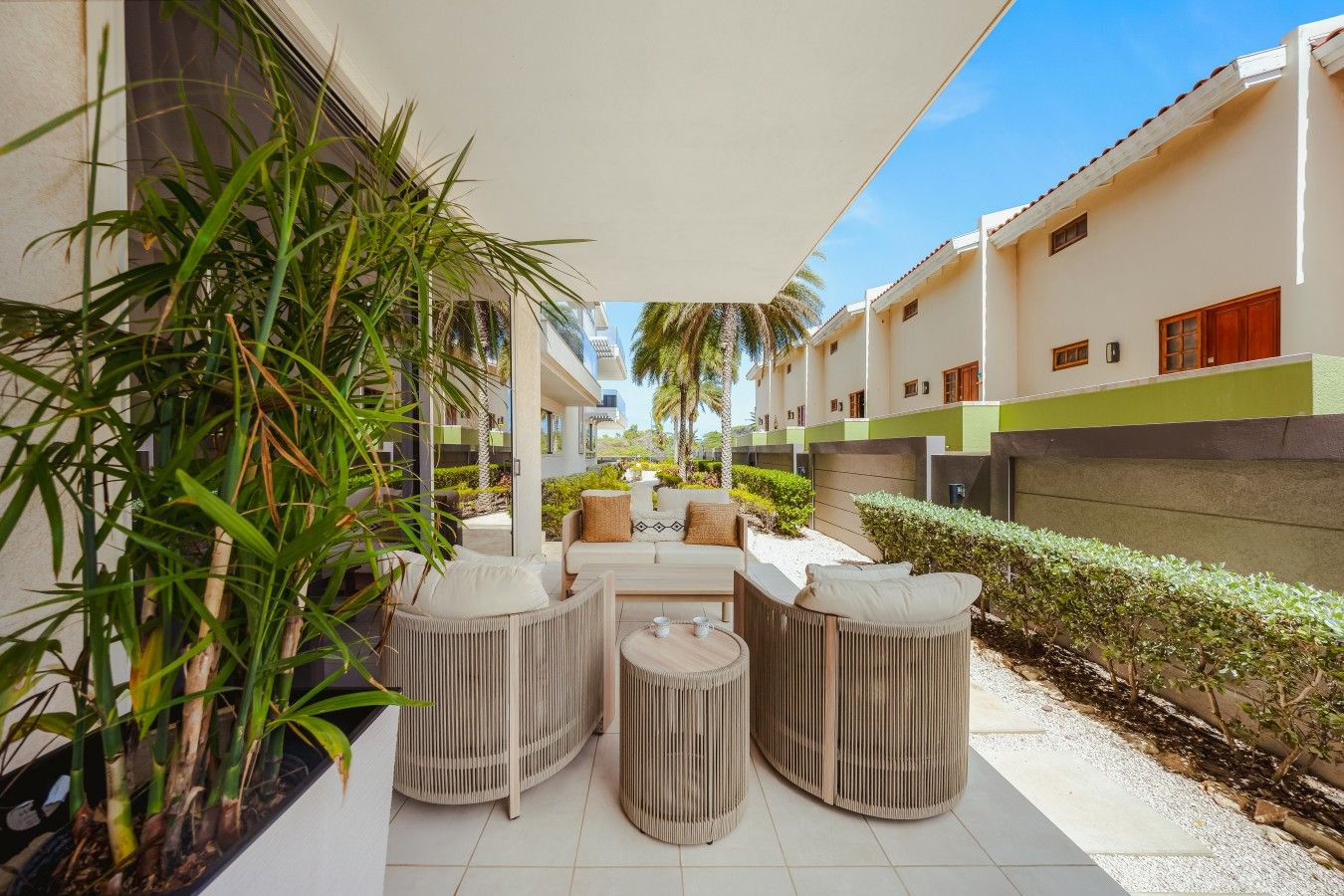 A covered outdoor seating area with wicker furniture, surrounded by greenery and a clear sky.