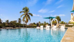 A clear blue pool with lounge chairs and umbrellas, surrounded by palm trees and a building with a curved balcony. - Coming Soon | O Condo 2 By Bocobay (Noord)