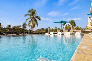 Une piscine d’un bleu éclatant, bordée de chaises longues et de parasols, entourée de palmiers et d’un bâtiment doté d’un balcon incurvé.