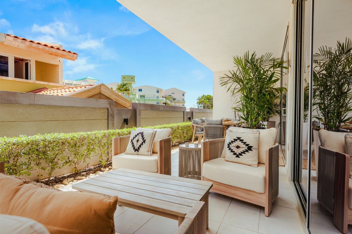 reverse-angle view of the bright outdoor terrace showcasing stylish armchairs with patterned cushions, a low wooden table, manicured green hedging along the privacy wall, and sunny blue skies above.