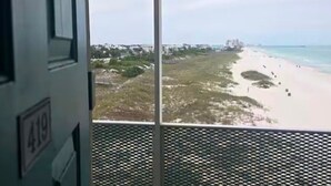 Une vue depuis une fenêtre ornée d’une grille métallique, donnant sur une plage où l’on aperçoit des personnes et des bâtiments au loin.