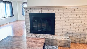Une pièce avec une cheminée, un plancher en bois, une fenêtre avec des rideaux et un luminaire au plafond.