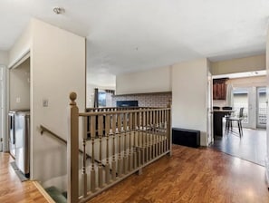 Amplia sala de estar con piso de madera, una escalera con barandilla y una cocina al fondo.