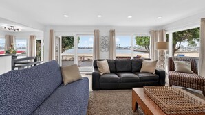 A living room with a black leather sofa, a patterned blue sofa, and a patterned armchair. There is a wooden coffee table and a decorative woven tray. The room has large windows with curtains, a view of the sea, and a balcony with a railing. - Newly updated 3-bedroom/3-bath beachfront house on the peninsula in Long Beach (Long Beach)