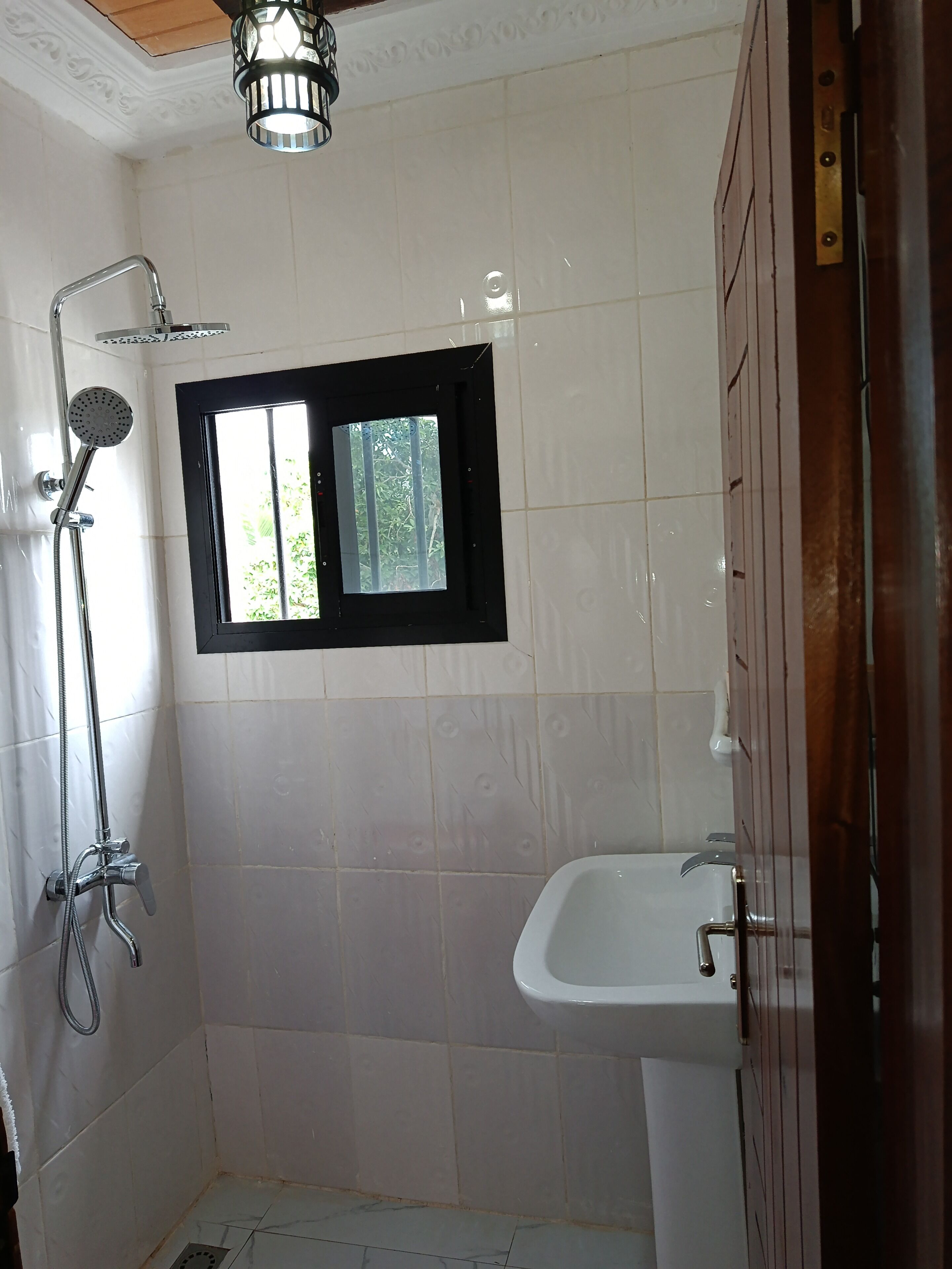 Ein Badezimmer mit weißen Fliesen, einer Dusche, einem Fenster und einem Waschbecken.