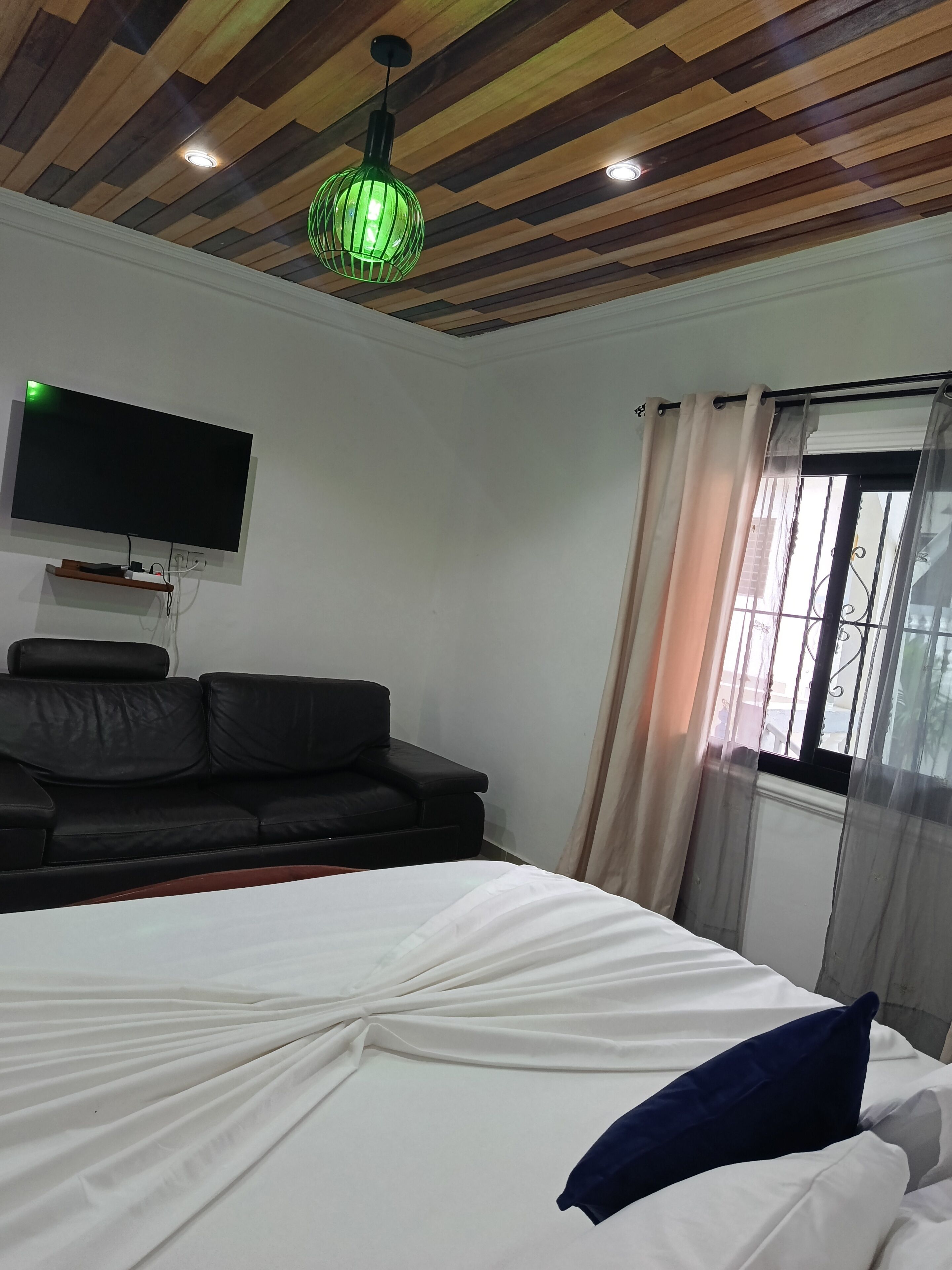 A room with a bed, a black sofa, a mounted TV, and a window with curtains.