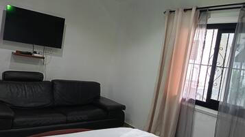 A room with a bed, a black sofa, a mounted TV, and a window with curtains.
