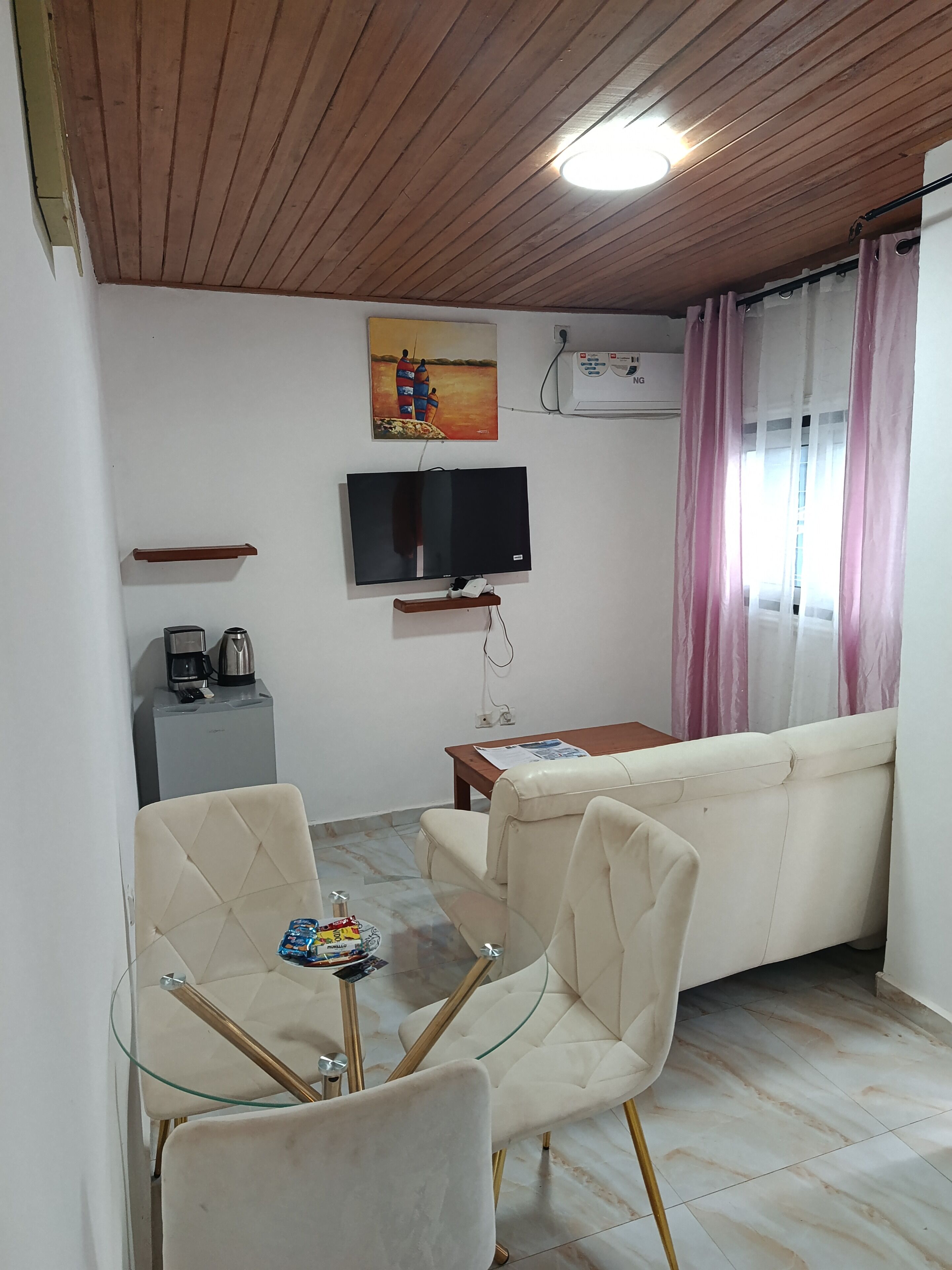 Una sala de estar con sofá, una mesa de vidrio y sillas, televisor, cafetera y un cuadro en la pared.