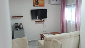 Una sala de estar con sofá, una mesa de vidrio y sillas, televisor, cafetera y un cuadro en la pared.