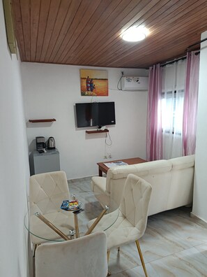 Una sala de estar con sofá, una mesa de vidrio y sillas, televisor, cafetera y un cuadro en la pared.