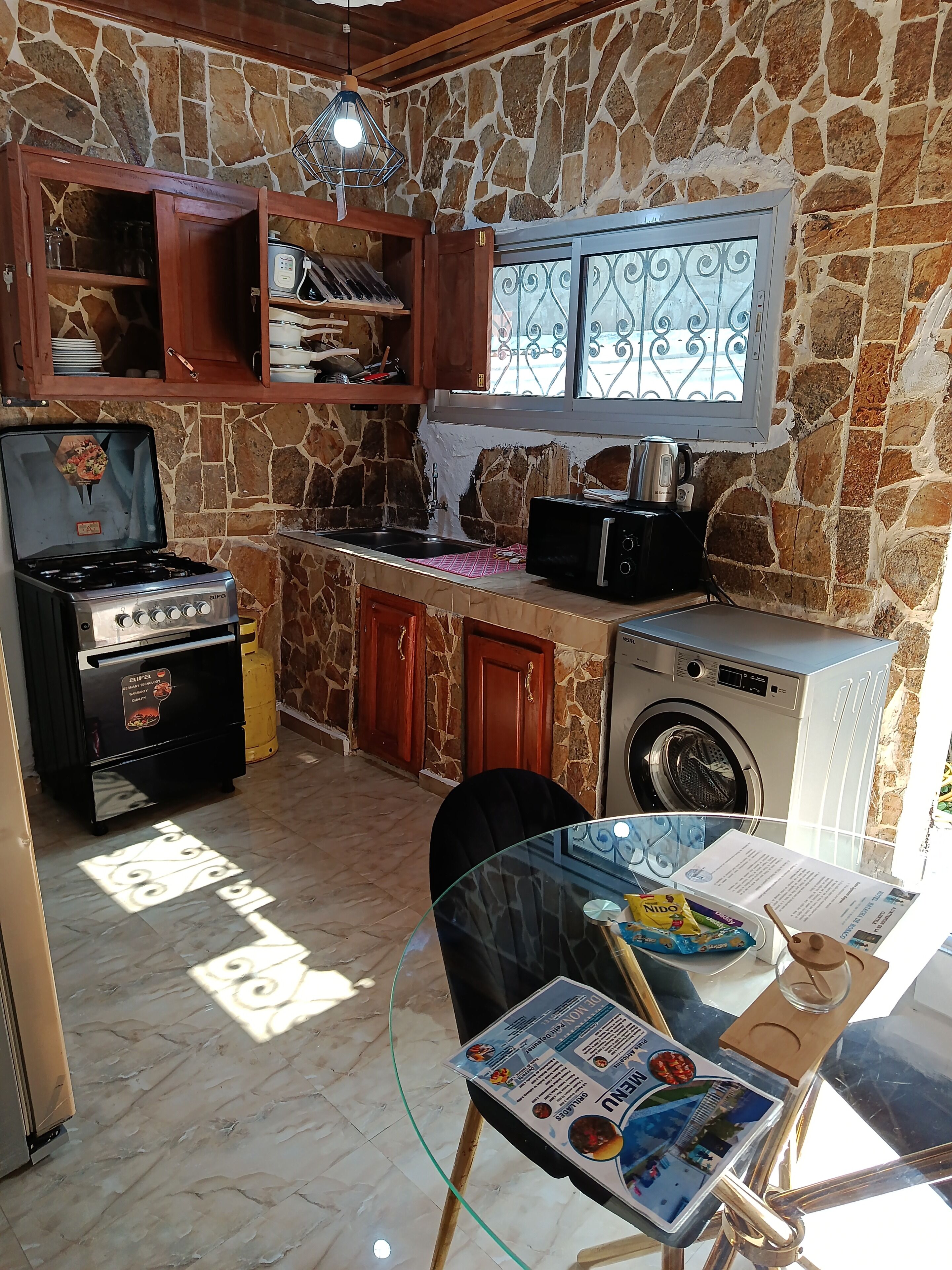 Une cuisine avec un mur en pierre, une cuisinière à gaz, un four à micro-ondes, un lave-linge et une table en verre avec un magazine.