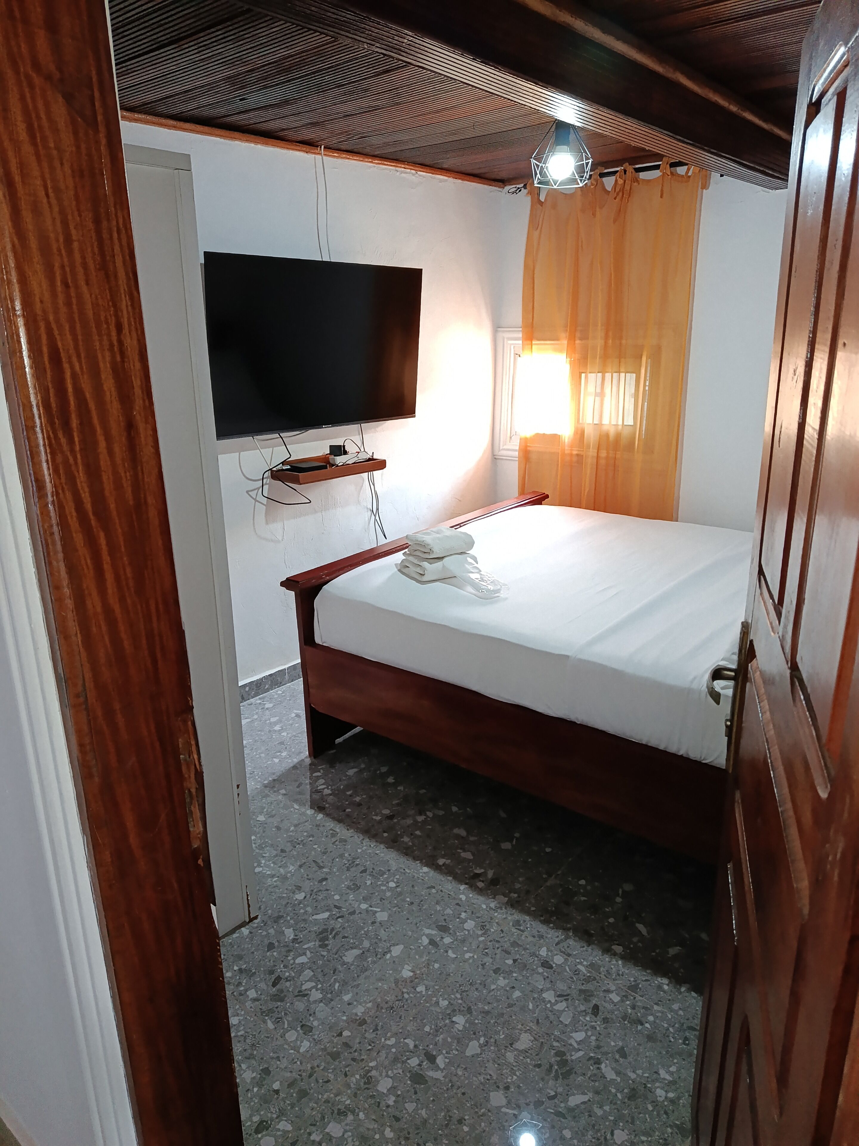 Une chambre d’hôtel avec un lit, un téléviseur à écran plat fixé au mur, une table de chevet avec une lampe et une fenêtre avec des rideaux.