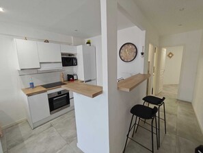 A modern kitchen with white cabinetry, a microwave, and a built-in oven. There is a wooden countertop and a black chair. - Appart 2 chambres – calme & lumineux – proche tram (Nice)